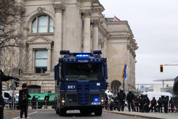 Gruppe von Polizeibeamten vor einem großen Gebäude mit Fahrzeugen auf der Straße und einer Person mit einer Kamera links, mit Bäumen, Ampeln, Flaggen und einem klaren blauen Himmel im Hintergrund.