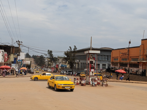 Ein gelbes Taxi fährt auf einer belebten Stadtstraße mit Gebäuden und Fußgängern, die Regenschirme halten, unter einem bewölkten Himmel, mit Strommasten und -kabeln im Hintergrund.