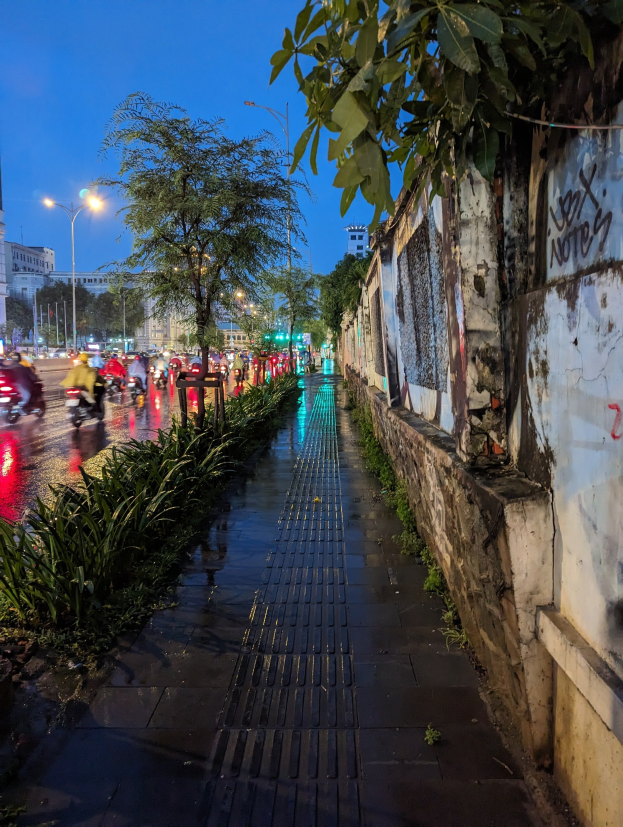 Stadtstraße bei Nacht mit Graffiti an der Seite, beleuchtet von Straßenlaternen, mit fahrenden Kraftfahrzeugen, Bäumen, Pflanzen, Gebäuden und einem sternenklaren Himmel im Hintergrund.