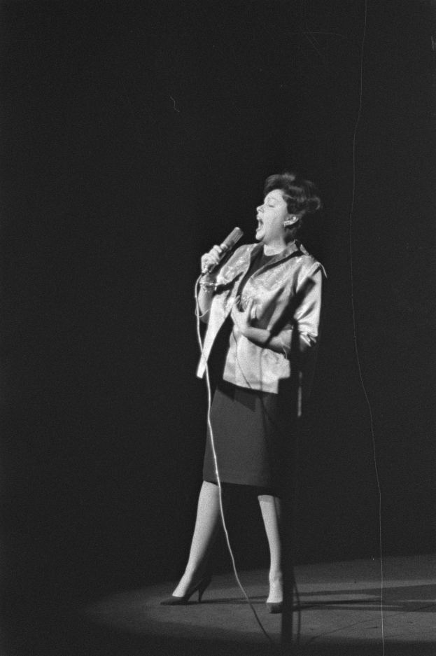 Eine Frau in einem schwarzen und weißen Outfit aus den 1950er-Jahren singt fröhlich auf der Bühne unter einem Scheinwerfer, hält ein Mikrofon.