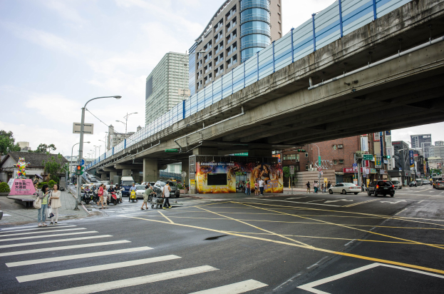 Eine belebte Stadtstraße mit einer Fußgängerbrücke darüber, Fahrzeuge, Fußgänger, Gebäude, Laternenpfähle und ein bewölkter Himmel.