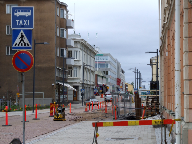 Eine Stadtstraße mit Gebäuden, Straßenlaternen, Schildern, Verkehrskegeln, Fahrzeugen, Absperrpollern, Bäumen und einem bewölkten Himmel.