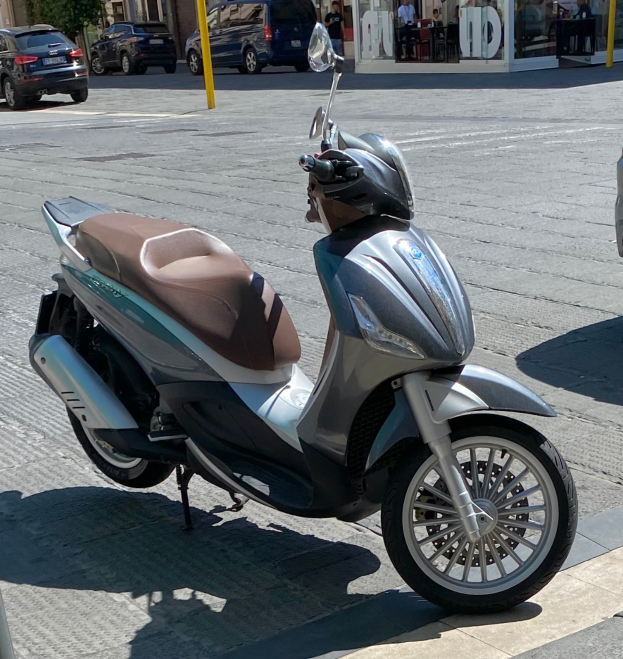 Ein Motorroller am Straßenrand mit einem Helm darauf, mit Gebäuden, Bäumen, Pfosten, Menschen, Stühlen, Tischen und anderen Gegenständen im Hintergrund.