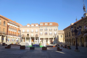Ein Plaza Mayor in einem Stadtplatz mit einem zentralen Brunnen, umgeben von Bänken, Topfpflanzen, Straßenlaternen, einer Uhrenturm und Gebäuden mit Fenstern unter einem klaren blauen Himmel.