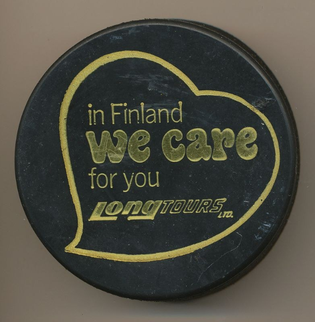 Schwarzer Hockey-Puck mit dem Text "In Finland We Care For You Long Tours" auf cremefarbenem Hintergrund.