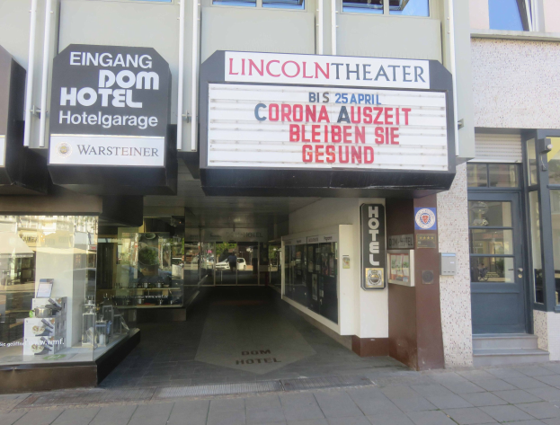 Außenansicht des Lincoln Theaters in Berlin, Deutschland, mit Glasfenstern und -türen und einer Tafel mit Text sowie einem Innenraum, der eine pulsierende Stadtkulisse suggeriert.
