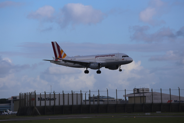 Germanwings Airbus A320-200 beim Start am Frankfurt Airport, mit Gras, einem Zaun, Gebäuden, Bäumen und einem bewölkten Himmel im Hintergrund.
