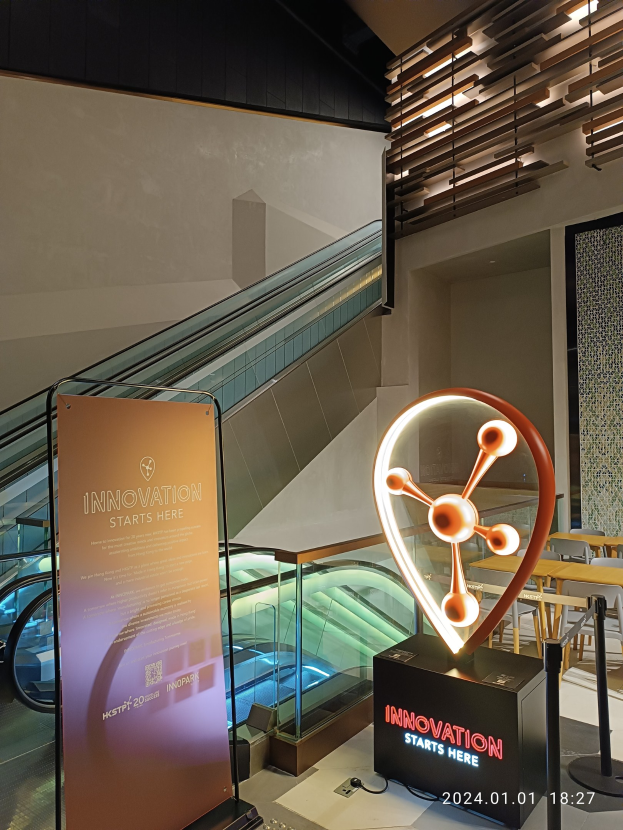 Großes Schild in der Mitte eines Gebäudes mit einer Rolltreppe im Hintergrund, Tische und Stühle rechts und eine Tafel mit "Innovation Starts Here" links, die auf die HSTP20 Expo hinweist.