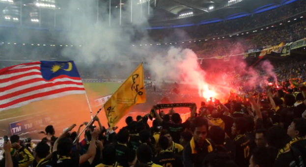 Große Menschenmenge in einem Stadion mit Fackeln, Fahnen und einer Kamera, mit Rauch und Schildern mit der Aufschrift "Malaysia vs Malaysia Live Streaming" im Hintergrund.