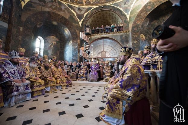 Eine Gruppe von Menschen in einer Kirche, einige sitzen und andere stehen auf einer Treppe, mit einer Person, die rechts eine Kamera hält; von der Decke hängt ein Kronleuchter, und an den Wänden hängen Gemälde.