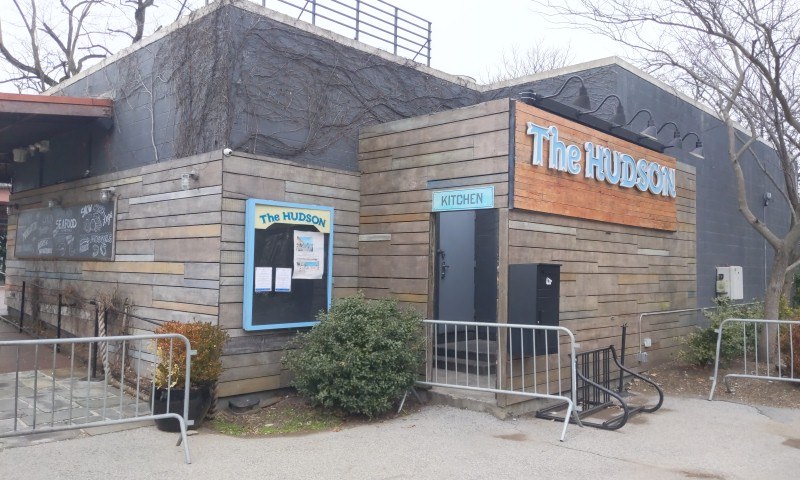 Außenansicht des Hudson-Restaurants in New York City, mit einer Tür, einem Schild, Topfpflanzen, einem Metallzaun, Bäumen, einem bewölkten Himmel und einem Müllcontainer im Vordergrund.