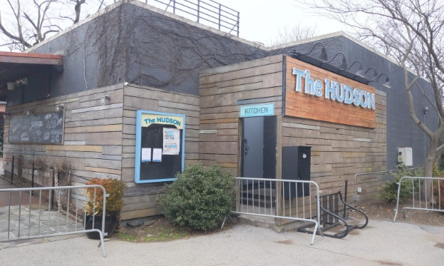 Außenansicht des Hudson-Restaurants in New York City, mit einer Tür, einem Schild, Topfpflanzen, einem Metallzaun, Bäumen, einem bewölkten Himmel und einem Müllcontainer im Vordergrund.
