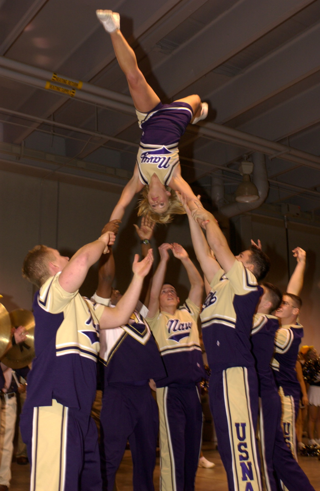 Eine Gruppe von Cheerleadern führt einen Stunt in einer Turnhalle durch, wobei eine Frau in einem Handstand in der Mitte steht und andere um sie herum stehen, einige halten links Musikinstrumente, und eine Wand mit Rohren und Deckenleuchten im Hintergrund.