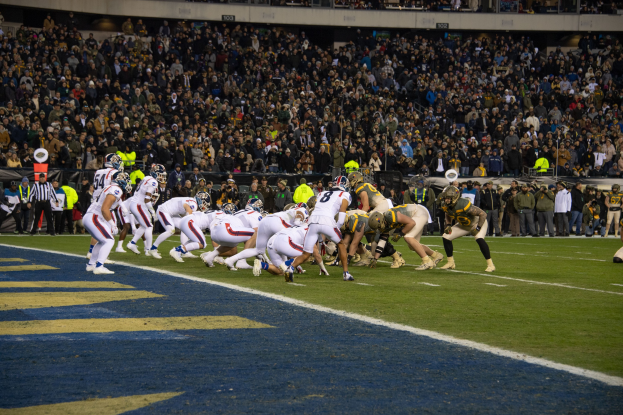 Ein Fußballspiel im Gange mit Spielern auf dem Boden in Sportkleidung und Helmen, betrachtet von einem Stadionplatz mit Zuschauern, Geländern und Beschriftungen im Hintergrund.