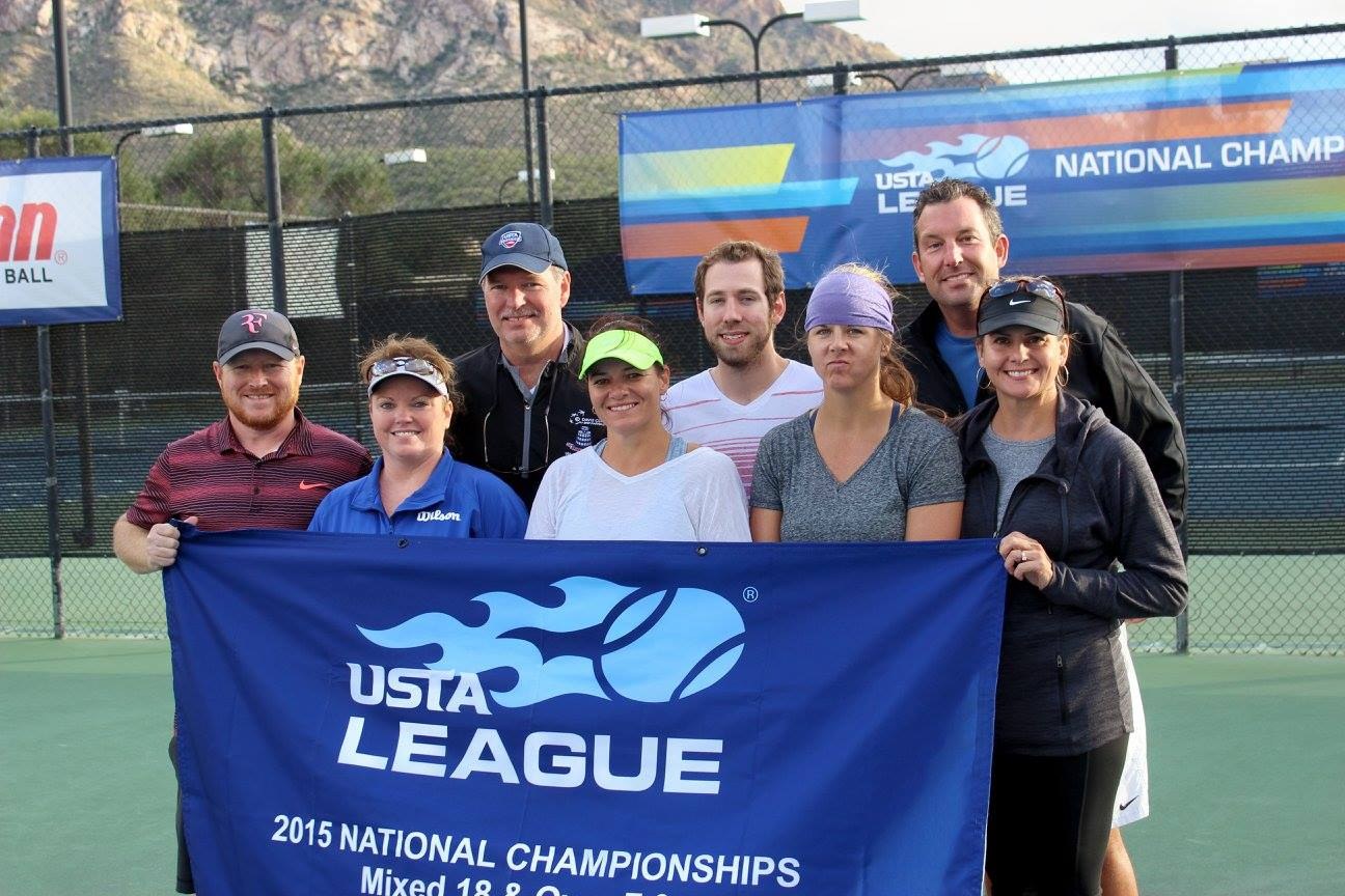 Gruppe von Menschen auf einem Tennisplatz mit einem "2015 USTA National Championships"-Banner, einem Zaun, Bannern, Laternenpfählen, Bäumen, Bergen und einem klaren blauen Himmel im Hintergrund.