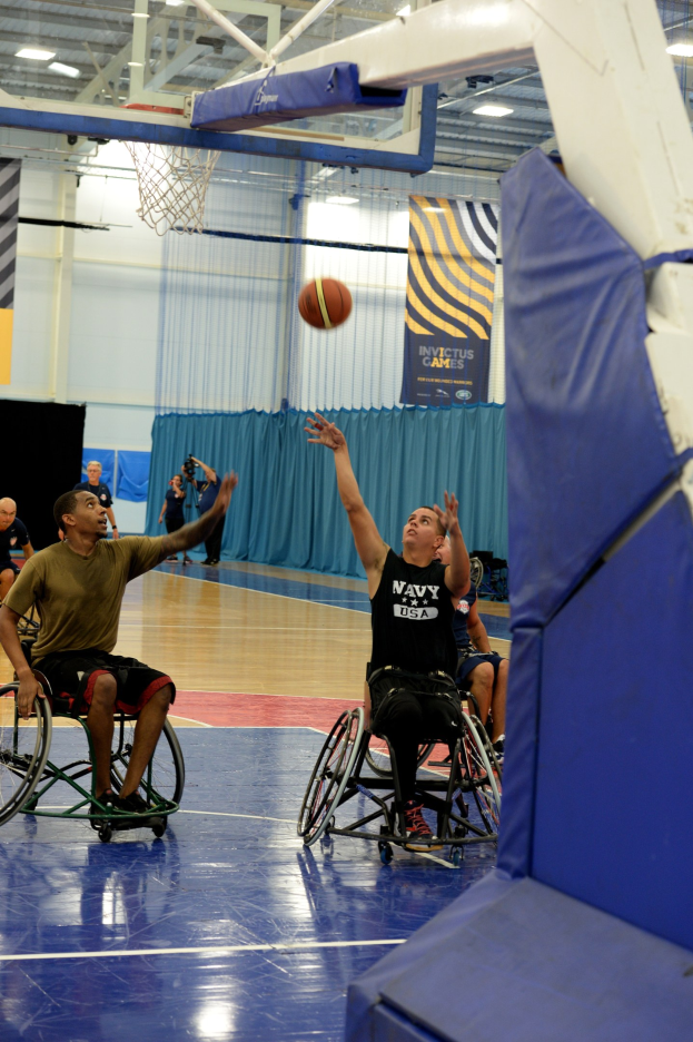 Eine Gruppe von Menschen im Rollstuhl spielt Basketball in einer Turnhalle mit einem Basketballkorb, Bannern mit Text und Deckenleuchten.