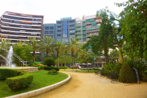 Ein Park mit einem zentralen Brunnen, umgeben von grünem Gras, Pflanzen, Bäumen, Bänken und Laternen, mit Fahrzeugen auf einer nahen Straße und hohen Gebäuden im Hintergrund unter einem klaren blauen Himmel.