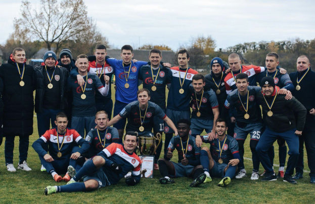 Männer in Sportbekleidung stehen auf einem Fußballfeld und halten Medaillen, mit einem Pokal zwischen ihnen, umgeben von Bäumen unter einem klaren blauen Himmel.