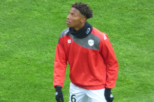 Ein junger Mann in einer roten Jacke und weißen Shorts steht auf einem Fußballfeld, trägt Baseballhandschuhe und schaut entschlossen nach rechts.