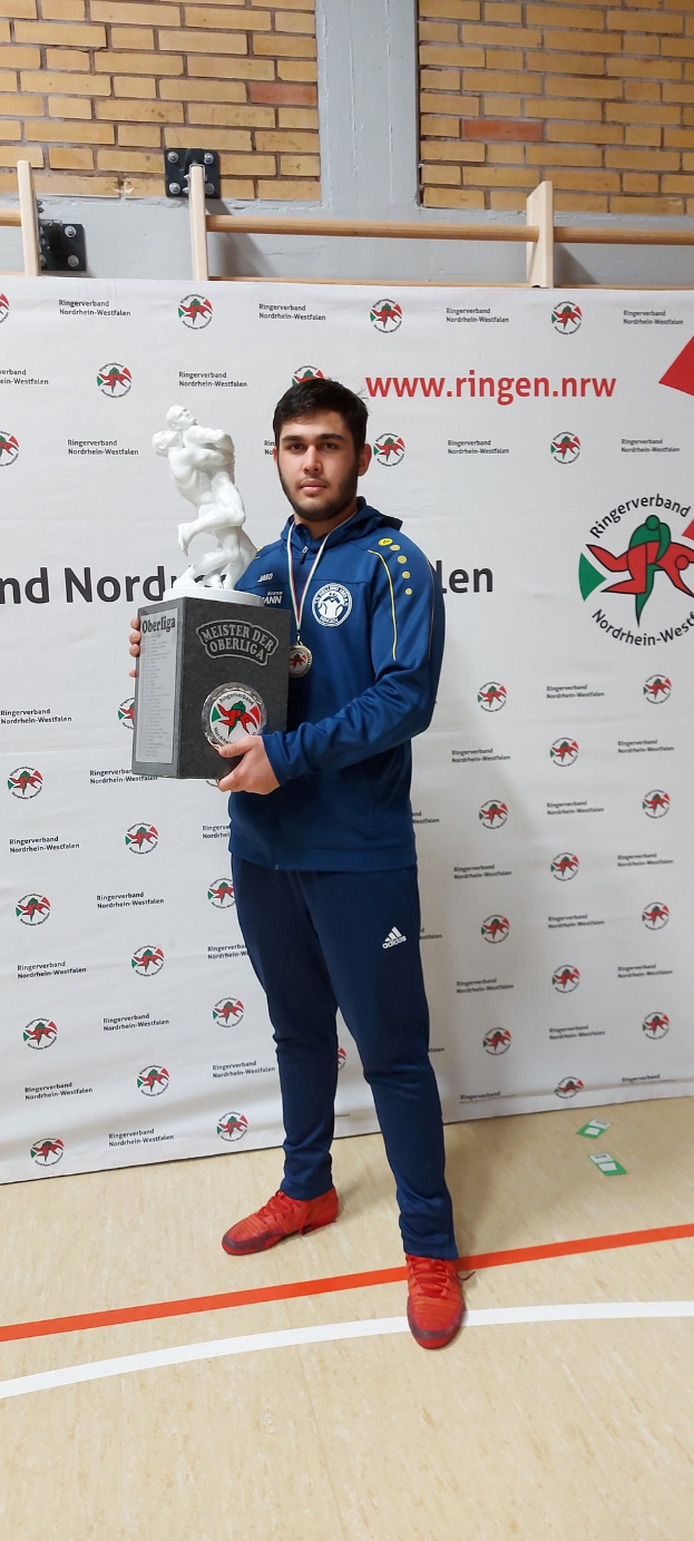Ein Mann in einem Anzug hält einen Pokal und trägt eine Medaille, während er vor einer bannergeschmückten Wand steht.
