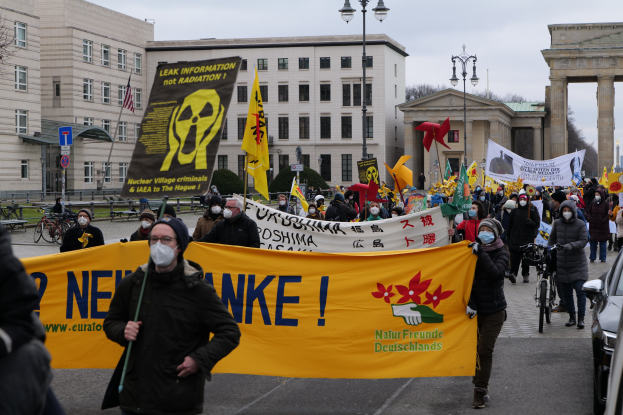 Große Gruppe von Menschen marschiert bei einer Straßendemonstration gegen Atomkraft in Deutschland, trägt Transparente und Fahnen, mit Fahrzeugen auf der rechten Seite und Gebäuden, Laternen, Bäumen und einem Tor im Hintergrund.