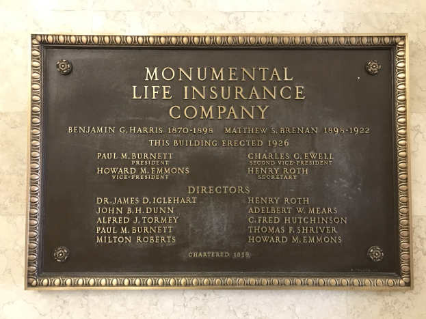 Plakette an einer Gebäudewand mit der Aufschrift "Monumentale Lebensversicherungs-AG" in fetter Schrift, innerhalb eines dekorativen Rahmens auf dunklem Material.