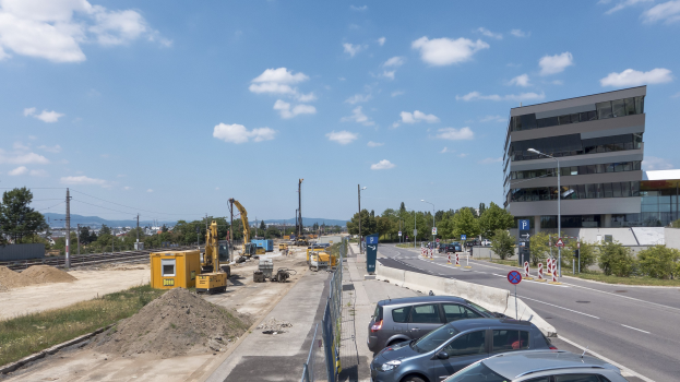 Eine Baustelle mit parkenden Autos, Gebäuden, Straßenlaternen, Straßenschildern, Absperrpollern, Bäumen, Pflanzen, Gras, Sand, Kränen, Bahnschienen, Strommasten mit Drähten, Hügeln und einer bewölkten Himmel im Hintergrund.
