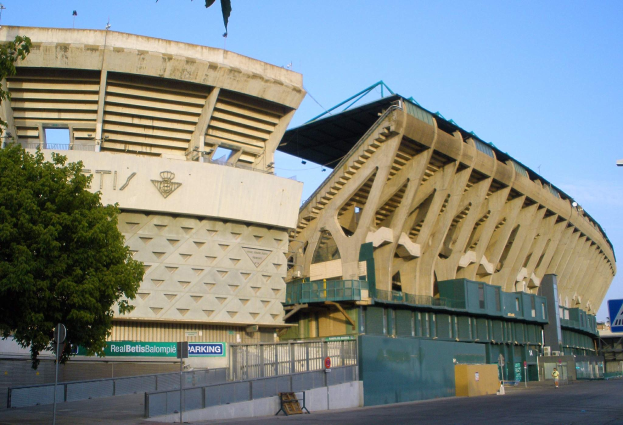 Großes Stadion mit zahlreichen parkenden Autos davor, umgeben von Gebäuden, Bäumen, Strommasten, Schildern, Geländern und einer Straße unter einem sichtbaren Himmel.