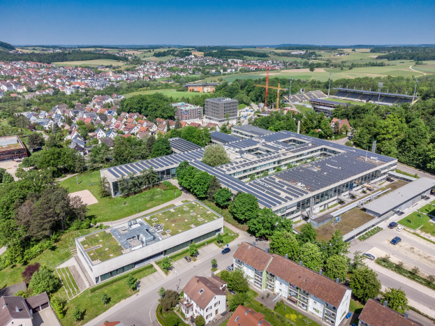 Luftaufnahme eines großen Gebäudes mit Solarmodulen auf dem Dach, umgeben von Häusern, Bäumen und Fahrzeugen auf einer Straße, mit Hügeln und einem klaren blauen Himmel im Hintergrund.