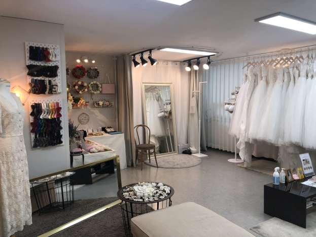 Ein unordentlicher Raum mit zahlreichen Hochzeitskleidern an Bügeln, einer Couch, einem Tisch mit Gegenständen, einem Stuhl, einem Spiegel, Vorhängen, Deckenleuchten und Wanddekorationen.