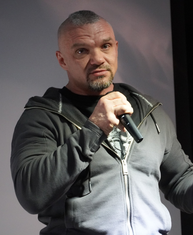 Ein Mann in einer schwarzen Kapuzenjacke und einem schwarzen Anzug mit weißem Hemd hält ein Mikrofon und steht selbstbewusst vor einer Wand, er scheint ein Komiker mit zurückgekämmtem Haar zu sein.