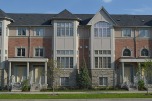 Eine Reihe von Stadthäusern in einer Wohngegend, mit Fenstern, Türen, Säulen, Geländern, Stufen, Pflanzen, Bäumen, Gras, einer Straße im Vordergrund und dem Himmel im Hintergrund.