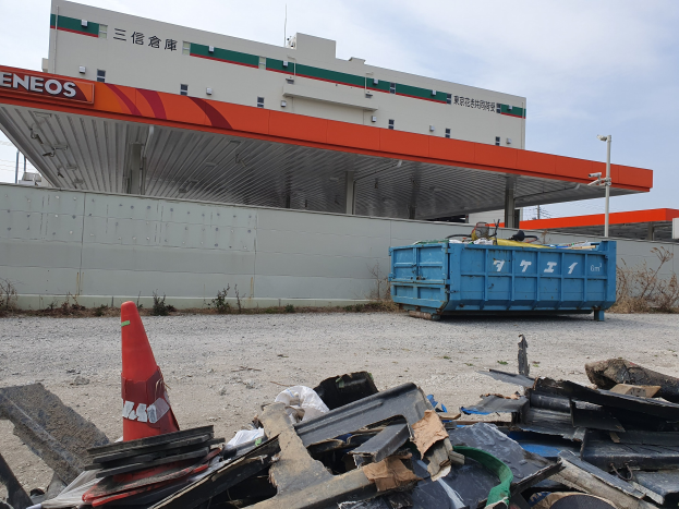 Großes Gebäude mit Haufen von Schrott, einem Verkehrskegel und einem blauen Container davor, umgeben von Pflanzen, Polen und Himmel während der Abrissarbeiten.