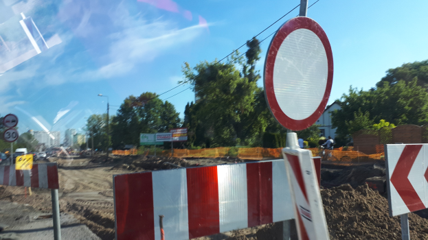 Baustelle mit einem roten und weißen Schild am Straßenrand, Bäume, Laternen, ein Verkehrszeichen im Vordergrund und Gebäude unter einem klaren blauen Himmel.