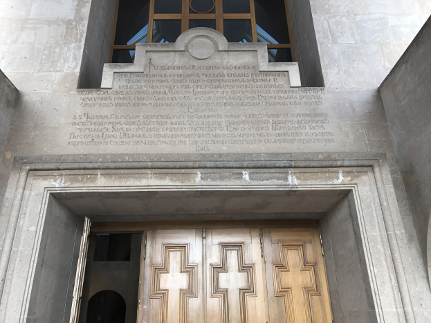 Große hölzerne Tür vor einem steinernen Gebäude mit einem Glasfenster oben, das likely eine Inschrift trägt, die Holocaust-Opfer gedenkt.