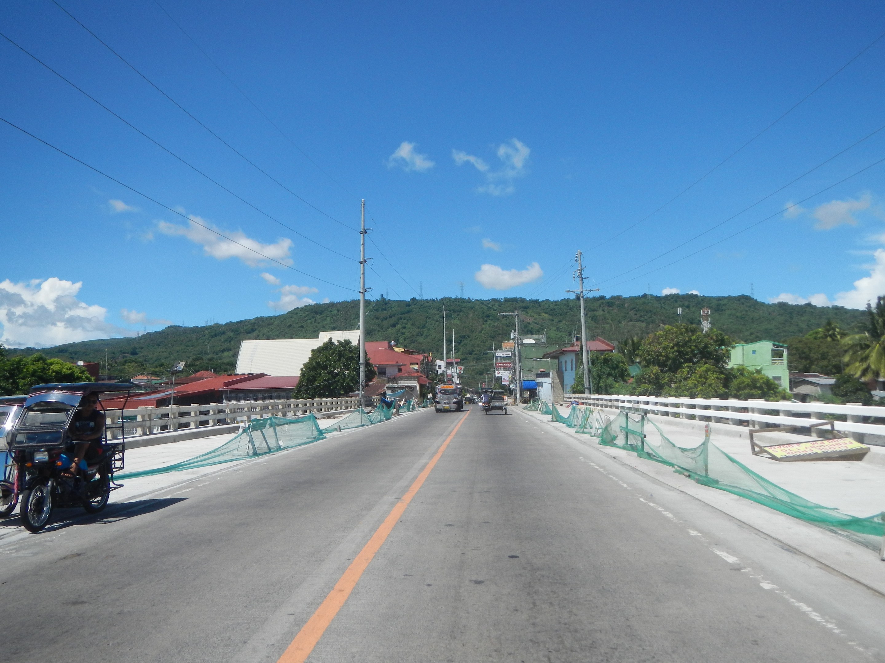Autobahn mit fahrenden Fahrzeugen, ein geparktes Motorrad auf dem Seitenstreifen, Geländer, Strommasten mit Drähten, Bäume, Gebäude, ferne Hügel und ein bewölkter Himmel.
