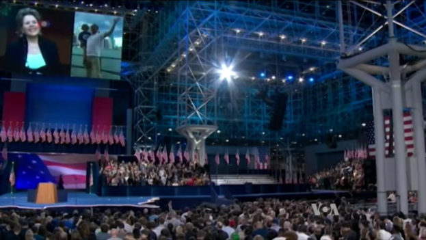 Eine große Menschenmenge sitzt vor einer Bühne mit einem Rednerpult, Mikrofon, Fahnen, einem Bildschirm und Beleuchtungsgeräten und wartet auf eine Präsidentschaftsdiskussion.