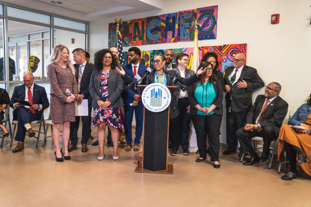 Gruppe von Menschen vor einem Rednerpult mit Mikrofon und einer Tafel mit der Aufschrift "Sancho-Stadtrat tritt dem Stadtrat bei", sowie verschiedenen Flaggen an der Rückwand.