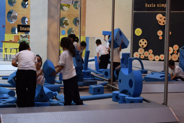 Kinder spielen in einer Turnhalle mit einigen, die Gegenstände halten, eine wandmontierte Tafel mit Text im Hintergrund und Stangen auf der rechten Seite.