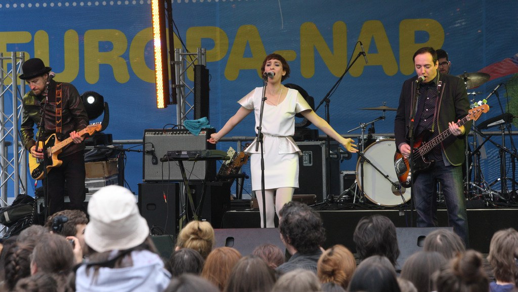 Vier Personen treten auf der Bühne auf: Eine Frau in einem weißen Kleid singt in der Mitte, flankiert von zwei Männern, die Gitarren spielen, während eine Menge zusieht, die von einer Wand mit Lautsprechern im Hintergrund umgeben ist.