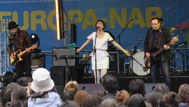 Vier Personen treten auf der Bühne auf: Eine Frau in einem weißen Kleid singt in der Mitte, flankiert von zwei Männern, die Gitarren spielen, während eine Menge zusieht, die von einer Wand mit Lautsprechern im Hintergrund umgeben ist.