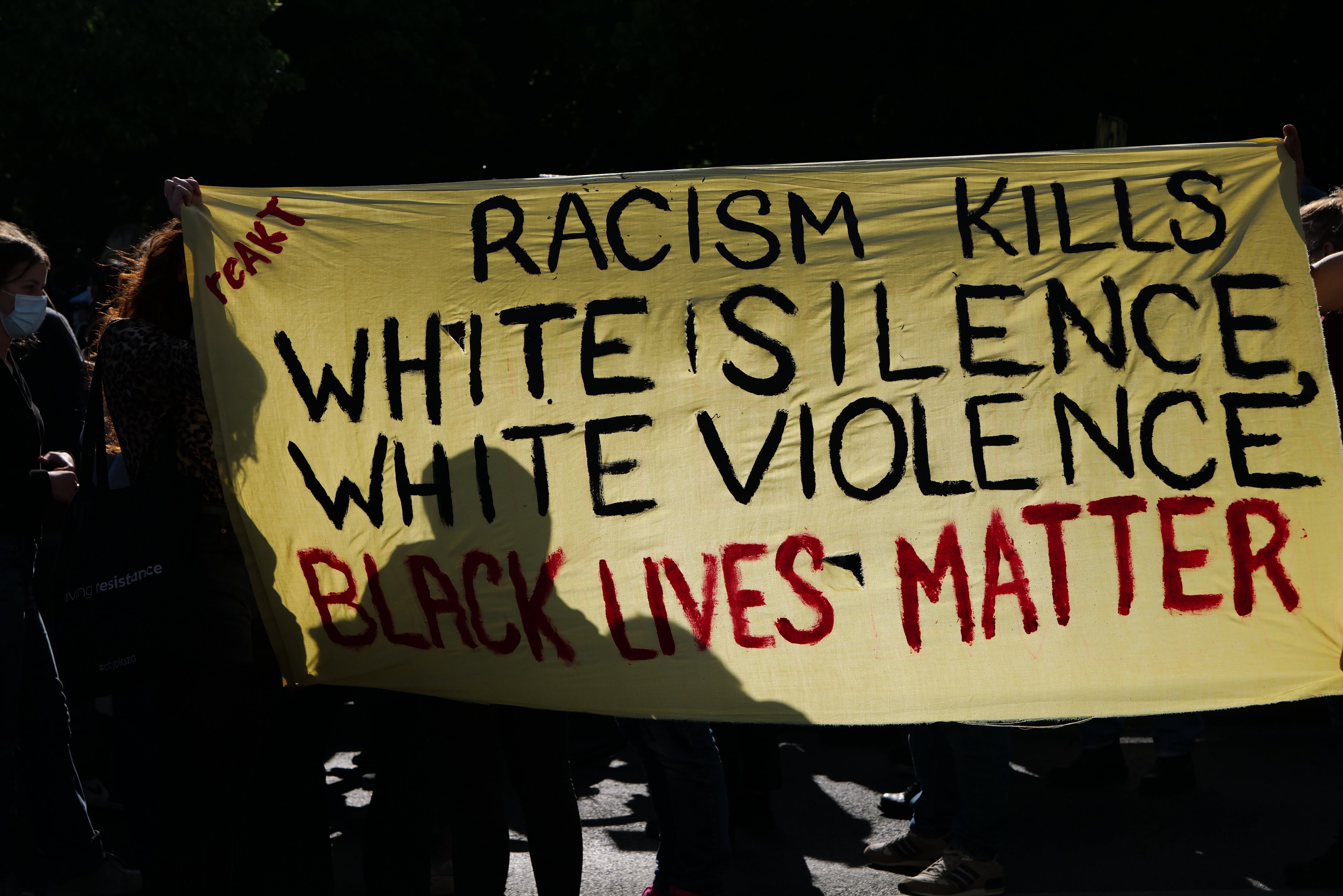 Eine Gruppe maskierter Personen hält ein Banner mit der Aufschrift "Rassismus tötet, weiße Stille, weiße Gewalt, Black Lives Matter" auf einer dunklen Straße.