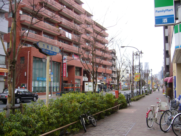 Eine Stadtstraße mit parkenden Fahrrädern, Gebäuden, Straßenlaternen, Straßenschildern, fahrenden Kraftfahrzeugen, Fußgängern auf dem Gehweg, Grünfläche und einem klaren blauen Himmel.