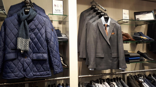 Kleiderbügeldisplay mit Anzügen, Krawatten, Jacken, Hemden und Schuhen, begleitet von Texttafeln und einer Wand im Hintergrund.