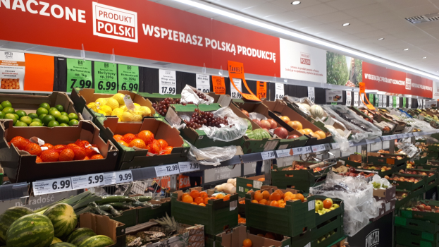 Ein Geschäftsraum mit verschiedenen Früchten und Gemüsen in Kartonschachteln mit Preisschildern, dazu Informationsschilder an den Wänden und Deckenbeleuchtung.