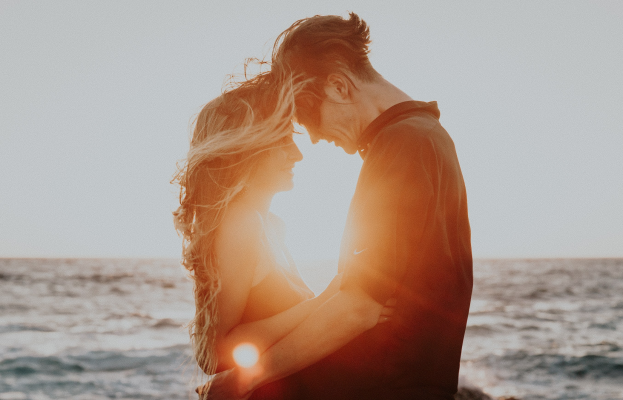 Ein Paar in Silhouette, das sich auf einem Strand bei Sonnenuntergang umarmt und küsst, mit leuchtend orangefarbenen, pinken und violetten Himmel und tiefblauem Wasser.