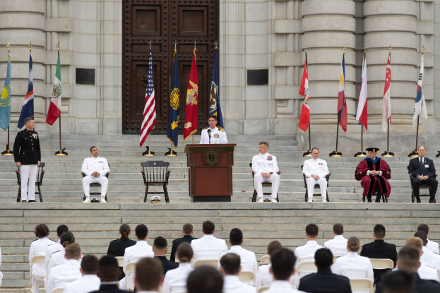 Eine Gruppe von Menschen in weißer Uniformen sitzt auf Stufen mit einem Podium, das von Fahnen flankiert ist, vor einem großen Gebäude mit einer Tür, was auf eine formelle Ehrung hindeutet.