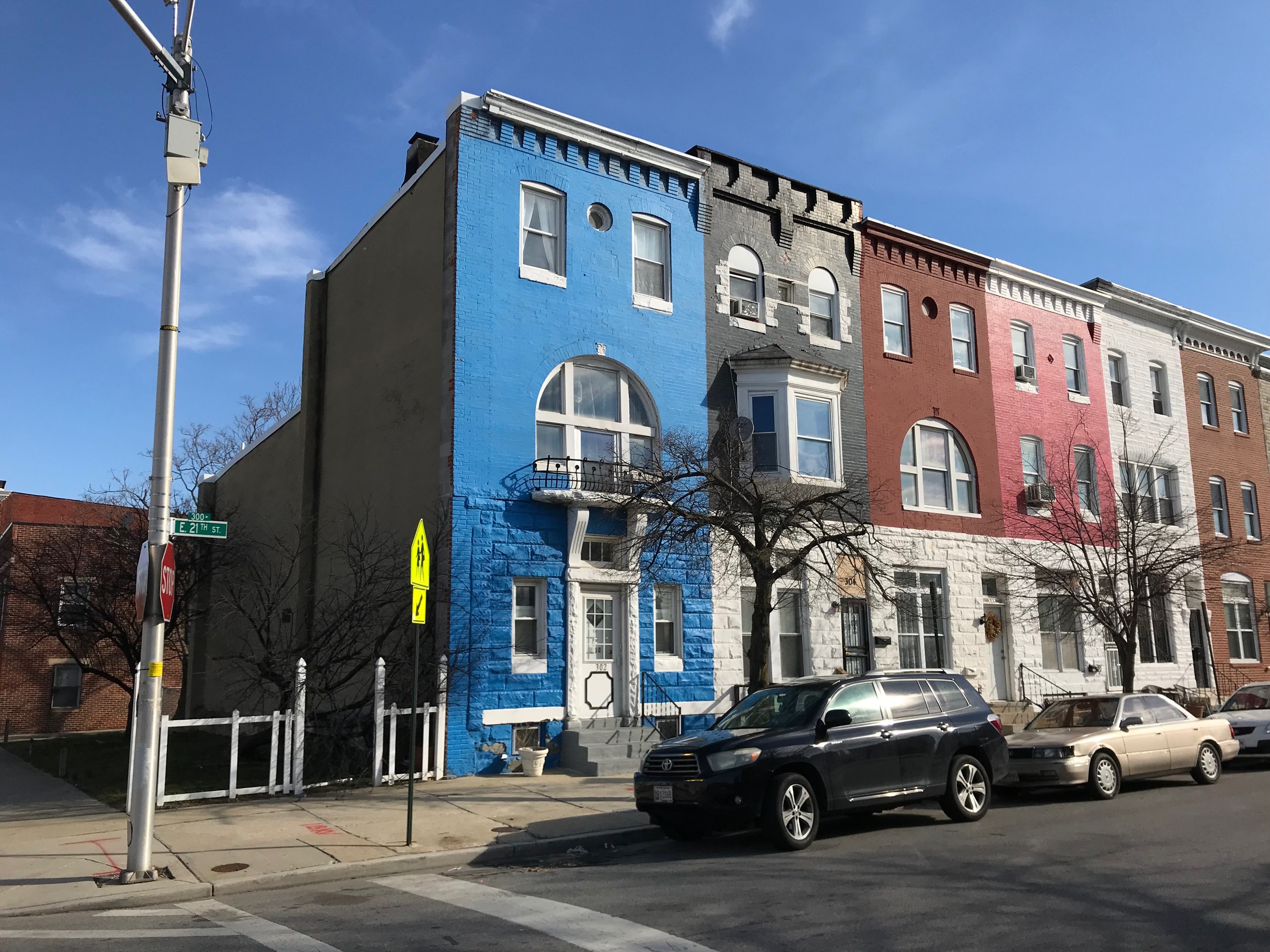 Eine Reihe bunter Häuser an einer Straßenecke mit parkenden Autos, Bäumen, Strommasten und einem Zaun unter einem bewölkten Himmel.