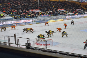Eine Gruppe von Menschen, die Hockey auf einer Indoor-Eisfläche spielen, umgeben von einem Zaun, mit Zuschauern im Hintergrund und beleuchteten Bannern darüber.