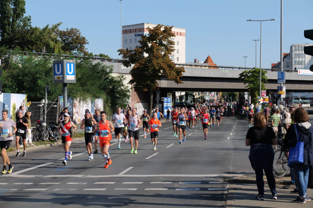 Gruppe von Menschen, die bei einem Marathon auf einer Straße mit Bäumen, Schildern, Fahrrädern, einem Zaun, Gras, einer Brücke, Gebäuden und einem klaren blauen Himmel laufen.